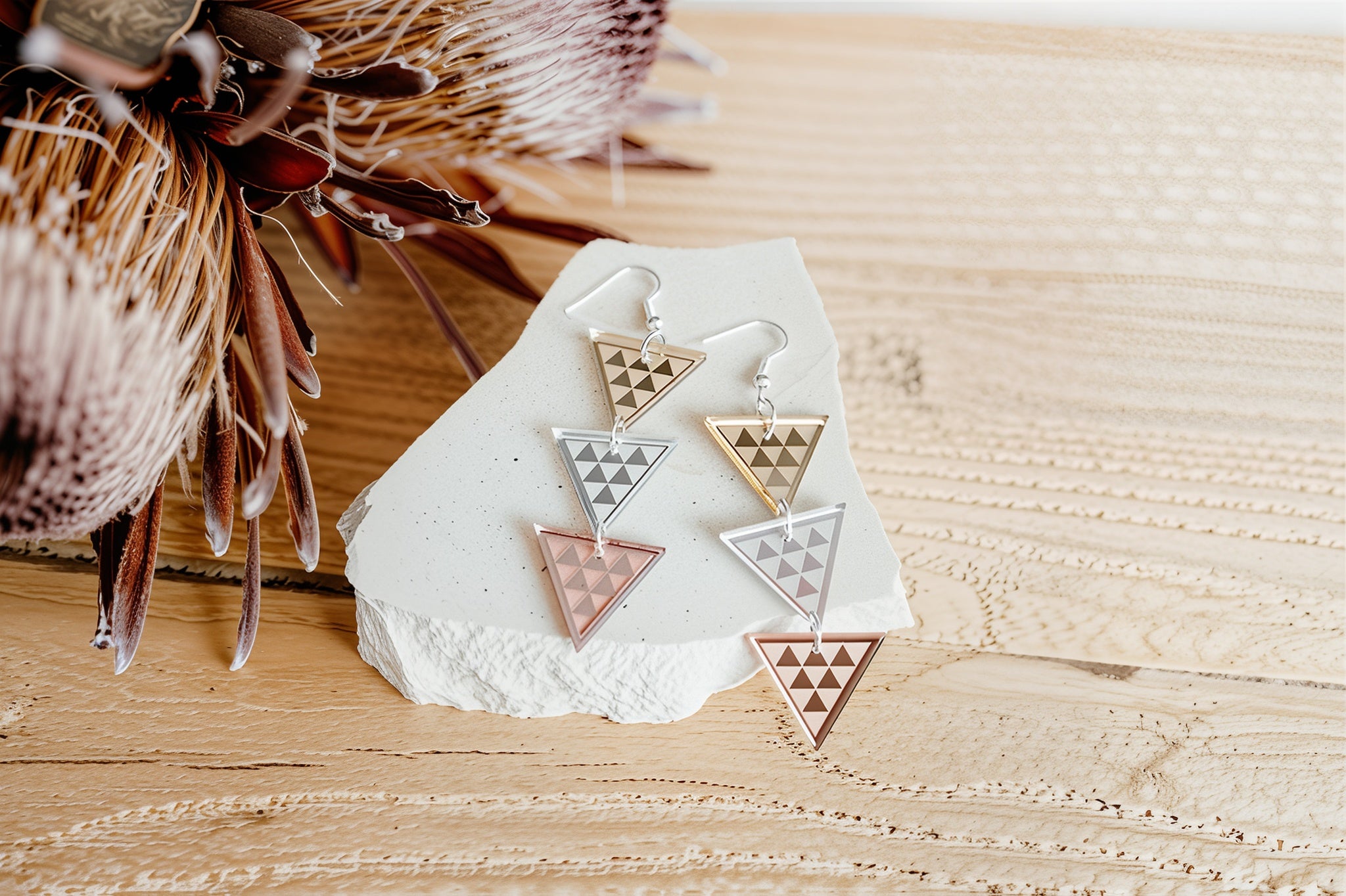 Lifestyle photo of Tāniko Triple triangle drop earrings in mixed metallics (silver, gold, rose gold) with engraved tāniko pattern, displayed on a white stone shard over light timber with a protea bloom.