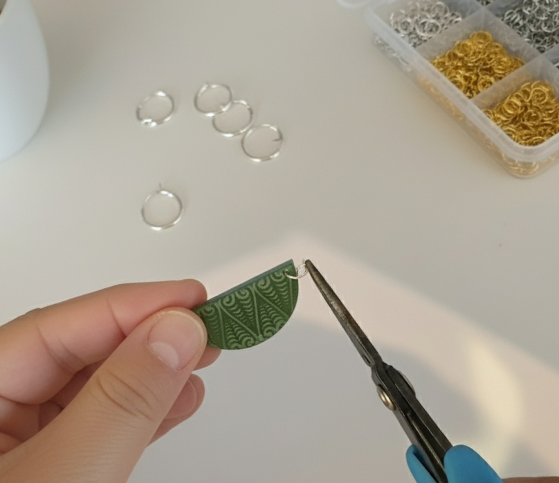 Close-up photo of hands assembling a green engraved Kōwhaiwhai earring using pliers, with silver hoops and findings laid out on a white workspace.