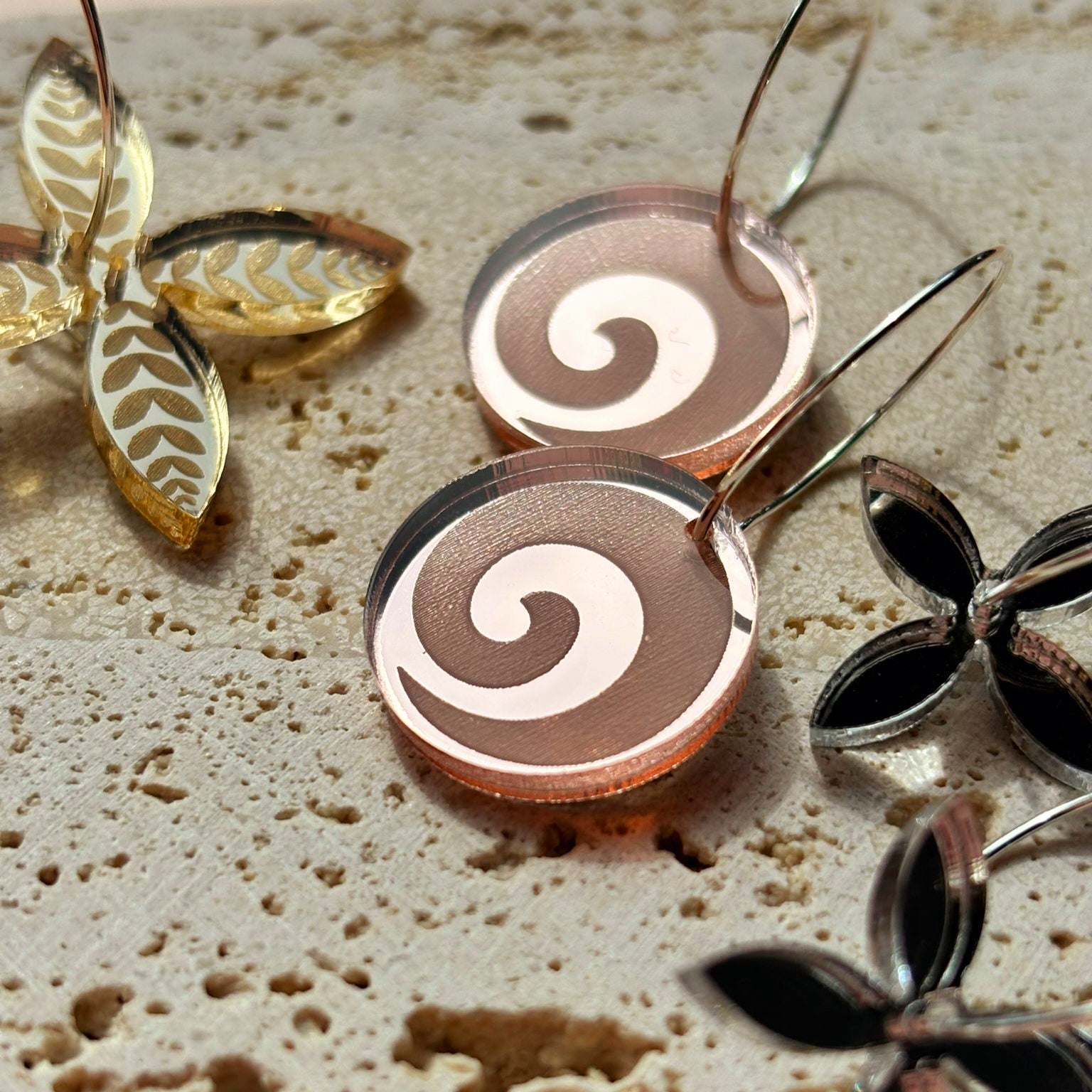 Koru Engraved hoop earrings on textured surface with decorative leaves; rose-gold mirror acrylic discs.
