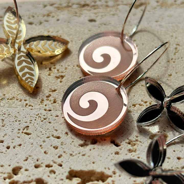 Koru Engraved hoop earrings on textured surface with decorative leaves; rose-gold mirror acrylic discs.