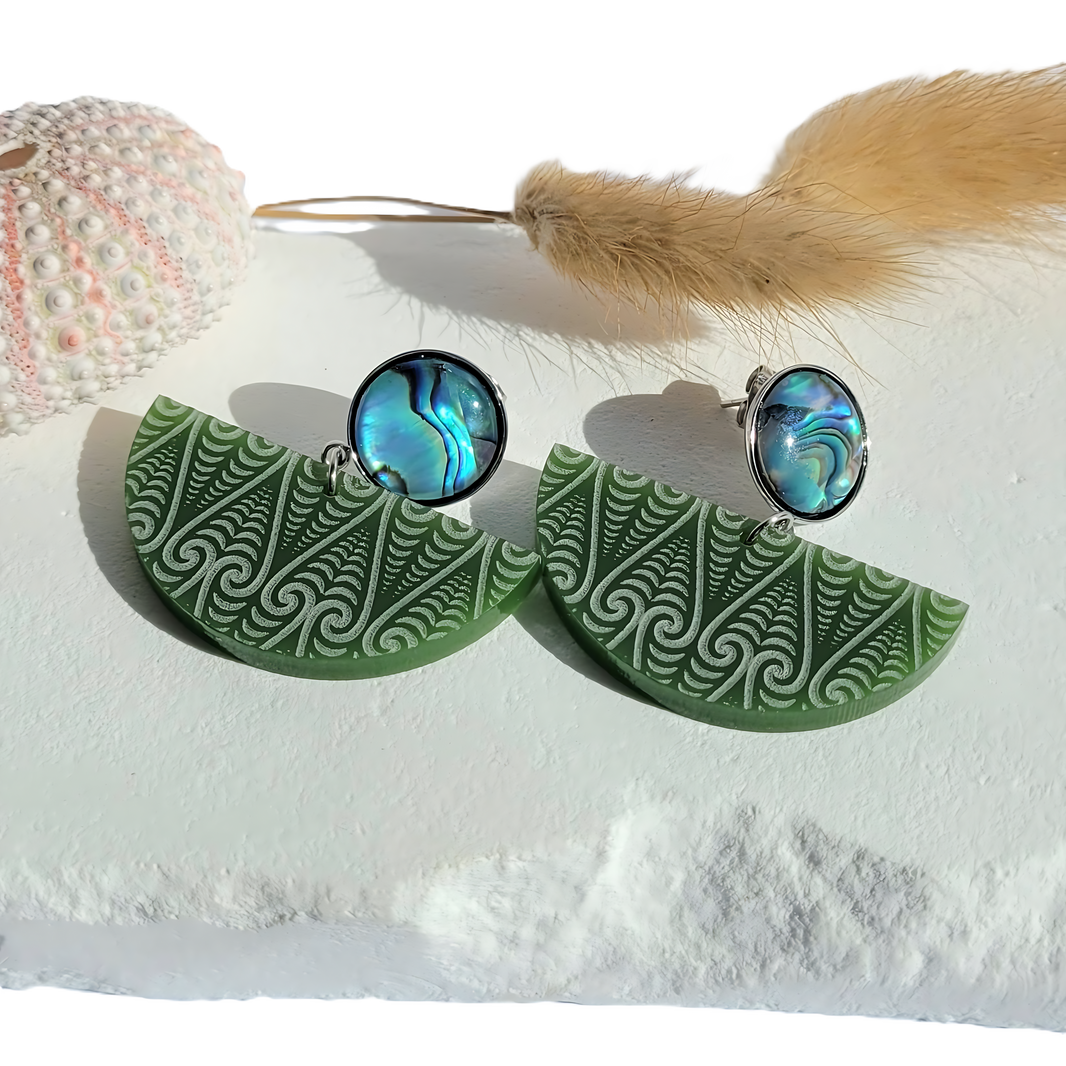 Lifestyle photo of Kōwhaiwhai Pāua stud earrings featuring engraved green acrylic kōwhaiwhai patterns with natural pāua shell tops, displayed on white stone with a sea urchin shell and dried grass.