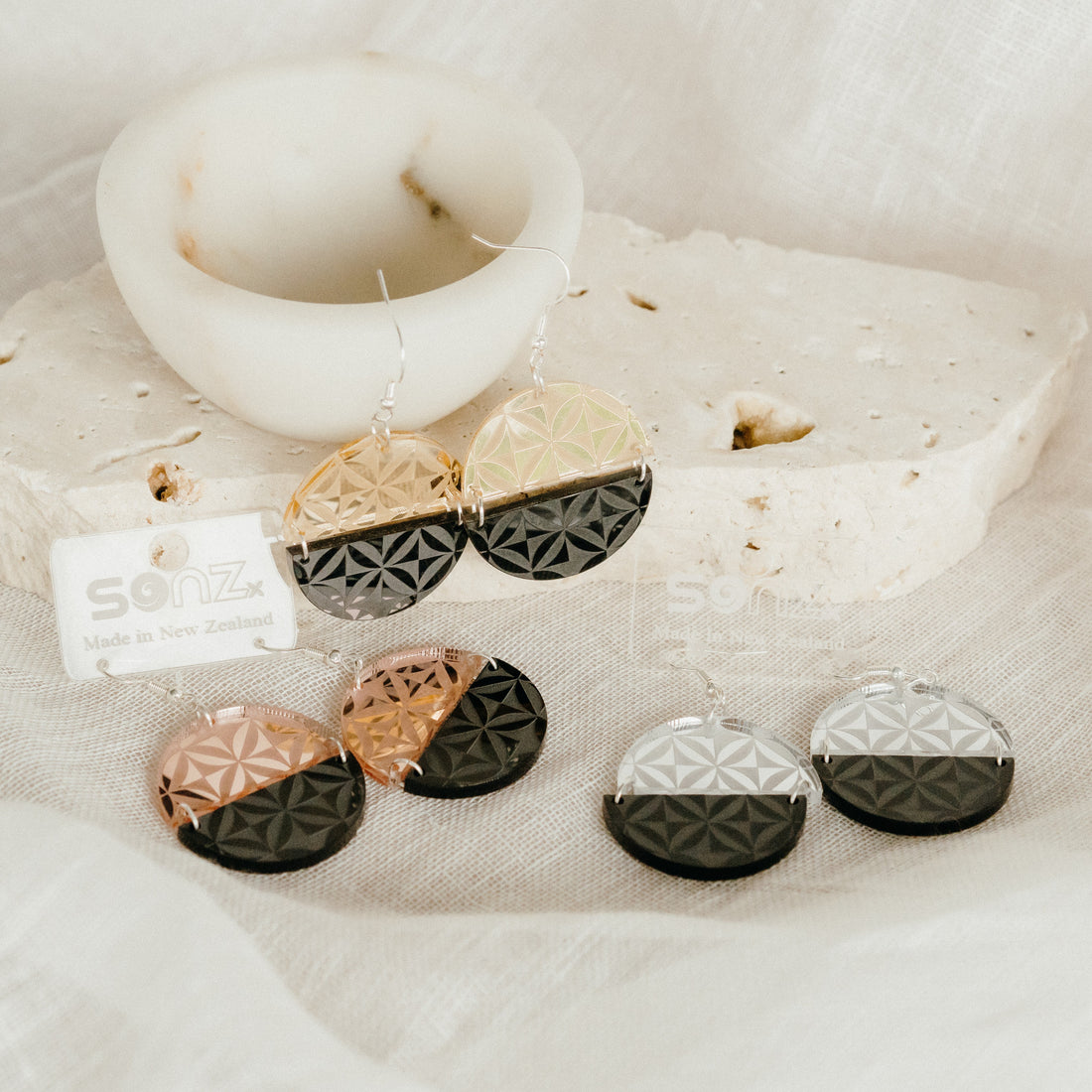 Manulua Symmetry earrings styled on stone with candle; gold mirror–black, rose-gold mirror–black and silver mirror–black pairs.