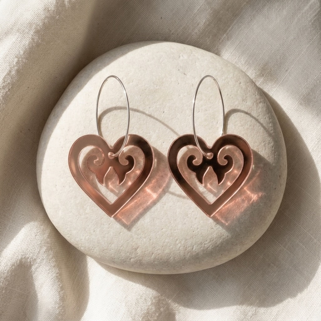 Aroha Heart Open earrings styled on a white stone; rose-gold mirror open hearts with koru scrollwork.
