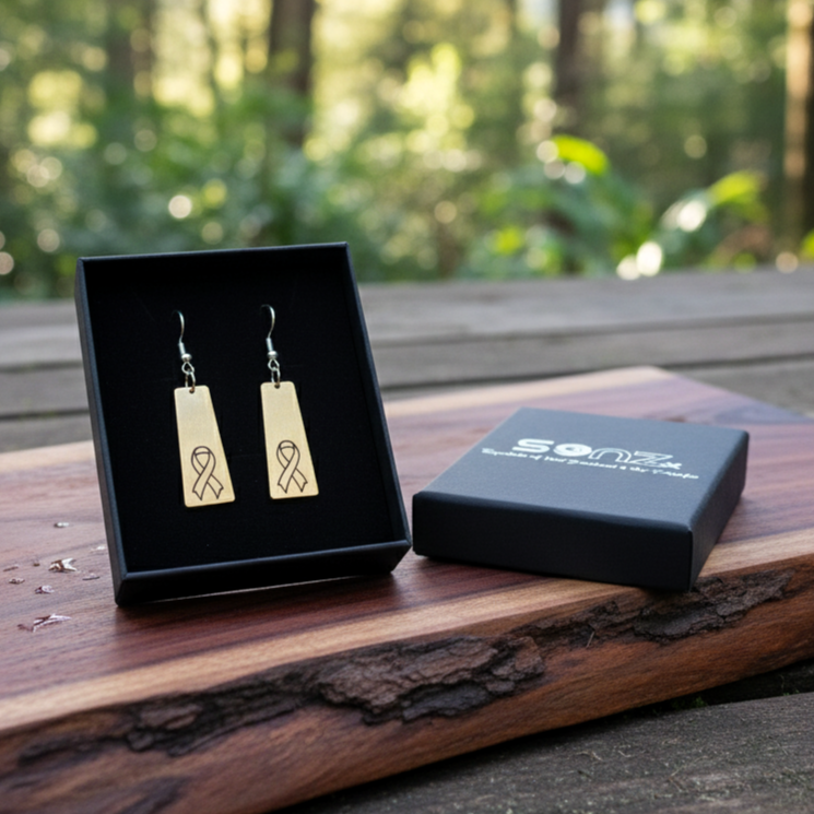 Lifestyle photo of Hugo’s Ribbon Bar earrings featuring engraved gold awareness ribbons on brushed gold acrylic, in SoNZ gift box on natural wood.