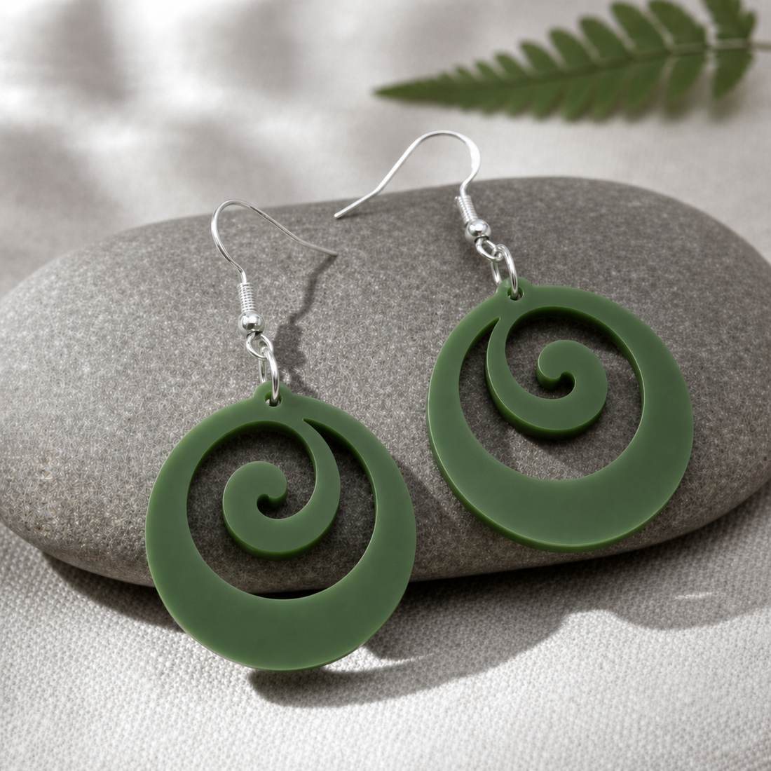Pair of mirrored Koru Flow earrings in fern-green Pounamu Satin Acrylic, arranged on a grey river stone over pale linen with a fern frond.