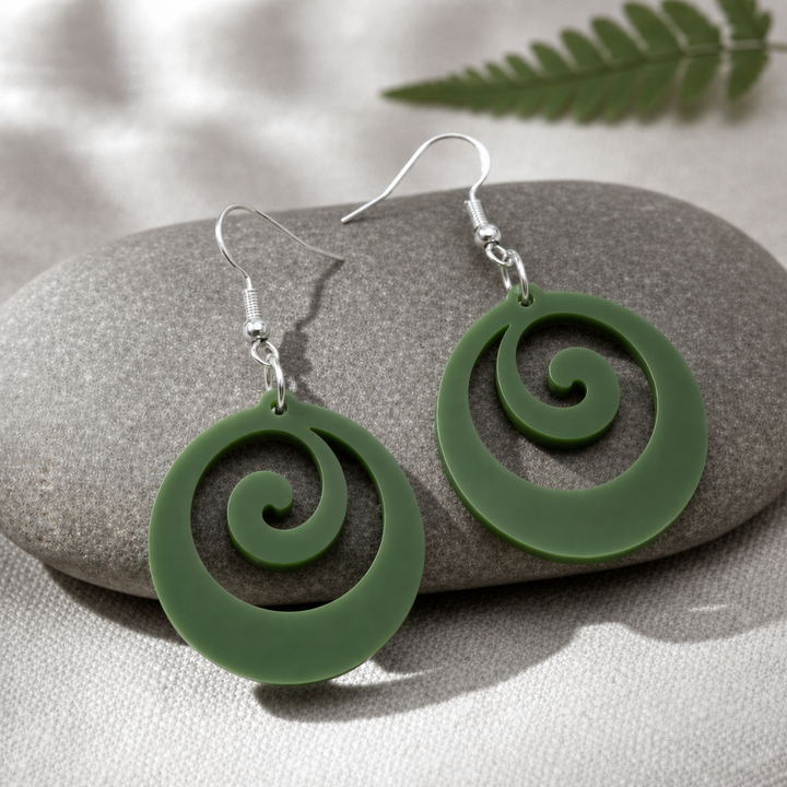 Pair of mirrored Koru Flow earrings in fern-green Pounamu Satin Acrylic, arranged on a grey river stone over pale linen with a fern frond.