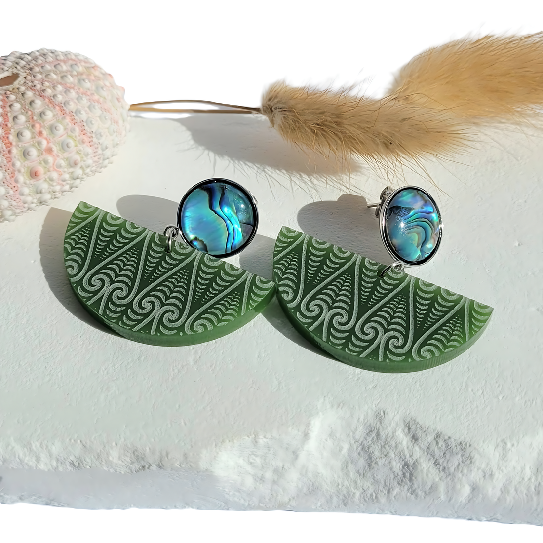 Kōwhaiwhai Pāua earrings on sand with shell and stems; pounamu satin half-moon and pāua stud.



