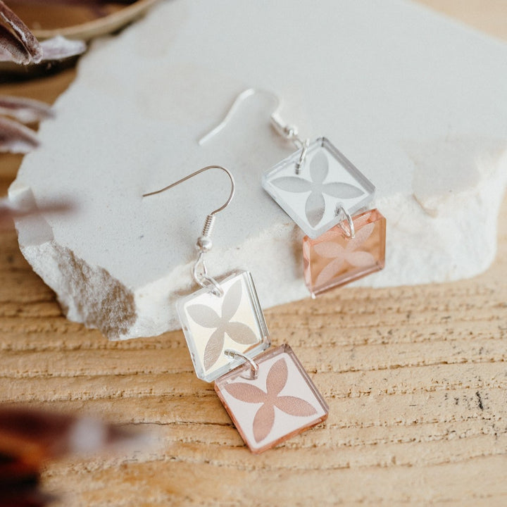 Tapa Double earrings on stone; stacked silver mirror and rose-gold mirror squares with tapa motif.